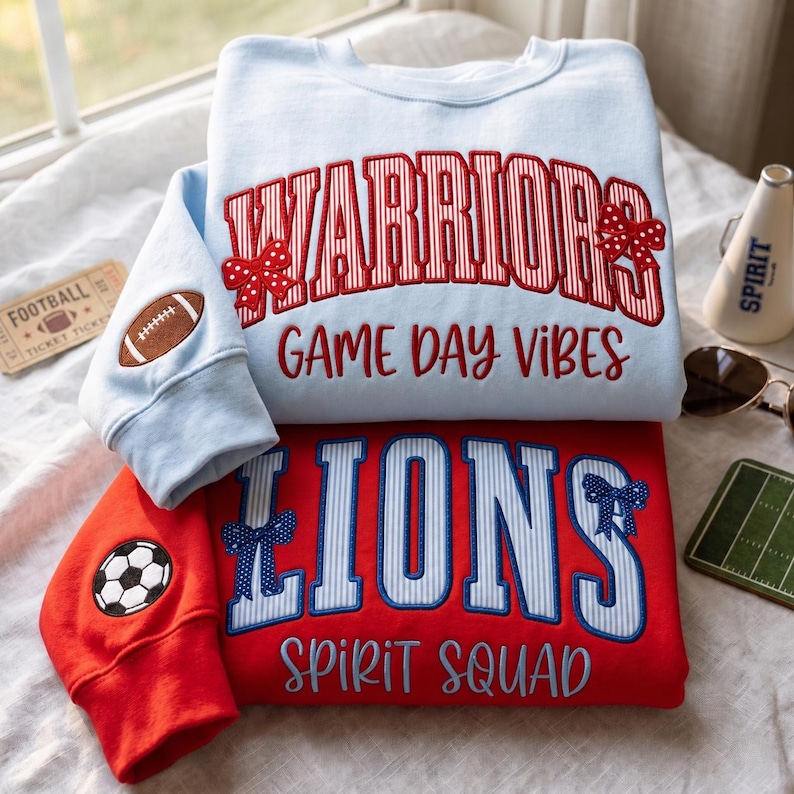 Op de afbeelding: Een lichtblauw sweatshirt met rood gestreepte WARRIORS-tekst, rode strikken en een football-patch op de mouw, over een rood sweatshirt met blauw gestreepte LIONS-tekst, blauwe strikken en een voetbal-patch op de mouw, beide met tekst.