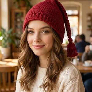 May include: A knitted red hat with a long, pointed tip and a tassel. The hat is made of a textured yarn and is worn by a person with long, wavy brown hair. The person is wearing a cream-colored sweater.
