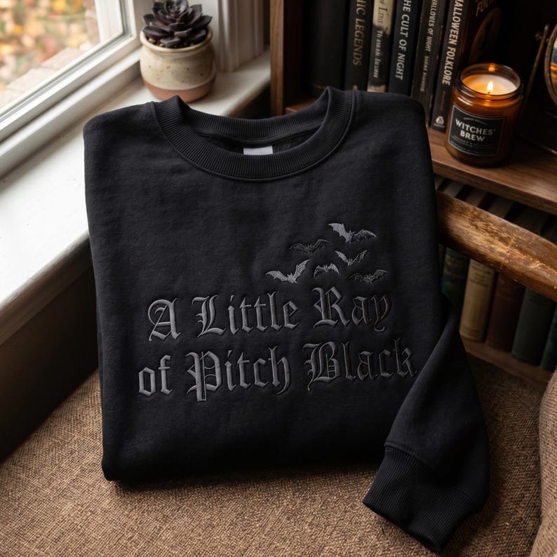 May include: Black crewneck sweatshirt with the phrase "A Little Ray of Pitch Black" embroidered in gray gothic lettering. Above the text are embroidered bats. The sweatshirt is displayed on a brown chair.