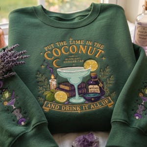 Könnte beinhalten: Ein waldgrüner Rundhals-Pullover mit detaillierter Stickerei eines Margaritaglases, Büchern, einer Kerze und Limetten. Der Text lautet "PUT THE LIME IN THE COCONUT", "MIDNIGHT MARGARITAS", "WITCHES SPIRITS" und "AND DRINK IT ALL UP". Die Ärmel sind mit passenden Motiven verziert.