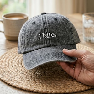May include: A gray baseball cap with the embroidered text "i bite." The cap is made of a faded denim material and has a curved brim. The cap is being held by a person.