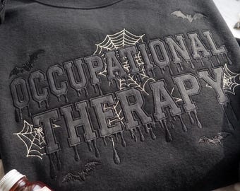 Może przedstawiać: Czarna bluza z wyhaftowanym napisem OCCUPATIONAL THERAPY w stylu kapiącej czcionki. Wzór zawiera białe, wyszywane pajęczyny oraz małe czarne nietoperze rozmieszczone wokół napisu na przedniej części odzieży.