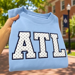 Peut inclure: Un sweat-shirt bleu clair à col rond arborant les lettres ATL dans une grande police de style bloc. Les lettres sont blanches avec un motif à pois noirs et sont soulignées par une bordure bleu marine foncé, créant un design audacieux.