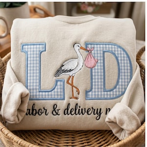 Könnte beinhalten: Beiges Sweatshirt mit blau karierten Buchstaben "LD" und einem Storch, der ein rosa Bündel trägt. Die Worte "labor & delivery" sind in schwarzer Schrift gestickt. Das Sweatshirt liegt in einem Weidenkorb.
