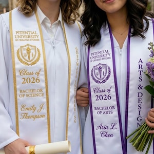 May include: Two people wearing white graduation gowns with gold and purple sashes. The sashes display the university name, degree, and class year 2026. One sash says "Bachelor of Science" and the other says "Bachelor of Arts".