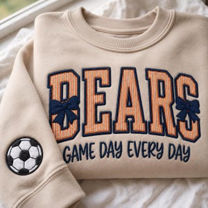 Op de afbeelding: Een beige sweater met ronde hals en de geborduurde tekst BEARS in oranje en marineblauw, versierd met marineblauwe strikjes op de B en S. Daaronder is de tekst GAME DAY EVERY DAY in marineblauw geborduurd. Een voetbal-patch bevindt zich op de mouw.