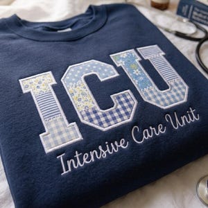 May include: A navy blue crewneck sweatshirt featuring large block letters that spell ICU. The letters are filled with a patchwork of light blue floral, striped, and gingham patterns. Below the letters, the text Intensive Care Unit is embroidered.