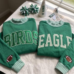 Puede incluir: Dos sudaderas verdes con cuello alto y puños de cuadros vichy blancos y verdes. Una sudadera muestra la palabra BIRDS en purpurina verde, la otra EAGLE en vichy. Ambas mangas presentan un parche marrón con forma de balón de fútbol americano.
