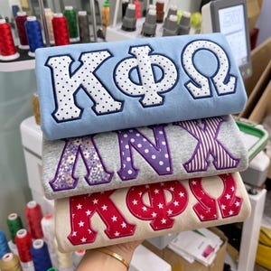 May include: Three folded sweatshirts stacked vertically. The top light blue sweatshirt features white Greek letters with black polka dots. The middle grey sweatshirt has purple letters with floral, polka dot, and striped patterns. The bottom cream sweatshirt has red letters with white stars.