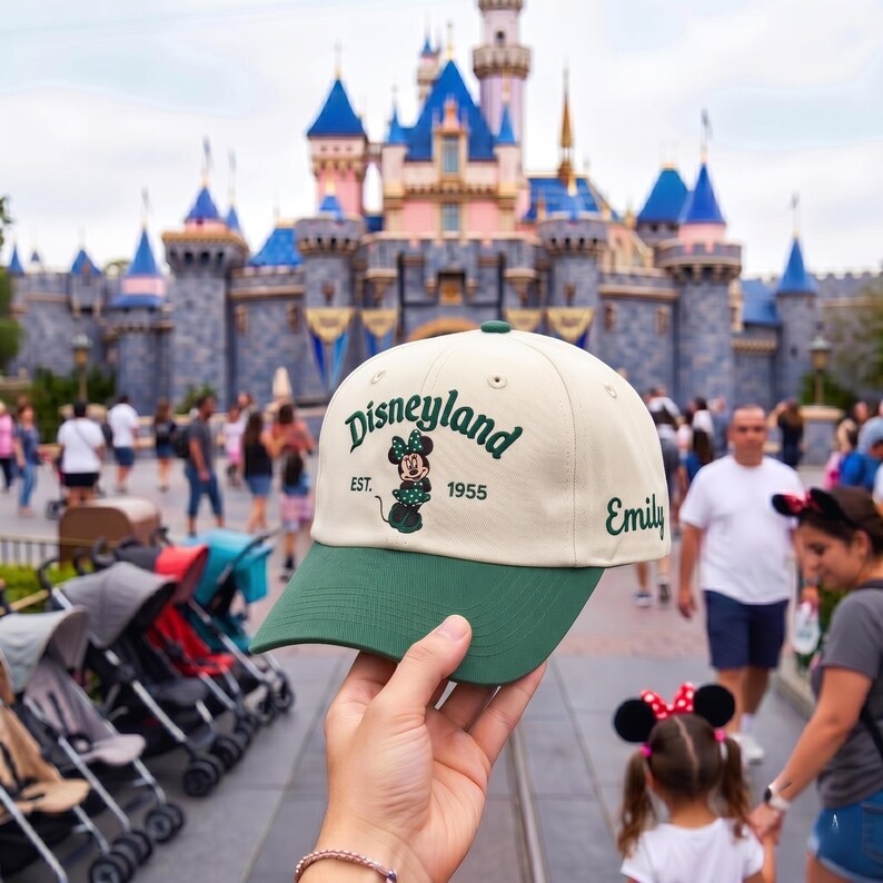 May include: A cream-colored baseball cap with a dark green brim. The front features the embroidered text Disneyland, EST. 1955, and a graphic of Minnie Mouse. The name Emily is embroidered in dark green cursive on the side of the cap.