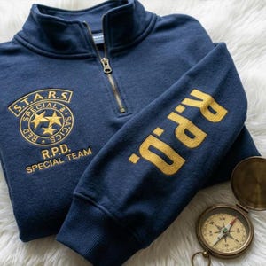 Könnte beinhalten: Marineblaues Sweatshirt mit Viertelreißverschluss und goldenen Stickereien. Der linke Ärmel zeigt "R.P.D." in Gold. Auf der Brust befindet sich ein goldener Aufnäher mit "S.T.A.R.S. SPECIAL & TACTICS" und "R.P.D. SPECIAL TEAM". Ein Kompass ist zu sehen.