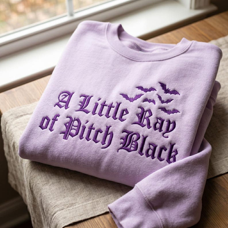 May include: A light purple sweatshirt with the embroidered phrase "A Little Ray of Pitch Black" in dark purple gothic lettering. Above the text are embroidered bats. The sweatshirt is folded on a wooden surface.