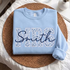 Könnte beinhalten: Ein hellblaues Sweatshirt mit Rundhalsausschnitt, das die Buchstaben MRS in einer weißen Blockschrift mit blauem Blumenmuster zeigt. Das Wort Smith ist in einer dunkelblauen Schreibschrift über die Mitte der Buchstaben gestickt.
