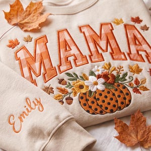 May include: A beige sweatshirt featuring the embroidered word MAMA in orange and white gingham letters. Below the text is an embroidered pumpkin with black polka dots, surrounded by autumn flowers and leaves. The name Emily is embroidered on the sleeve.