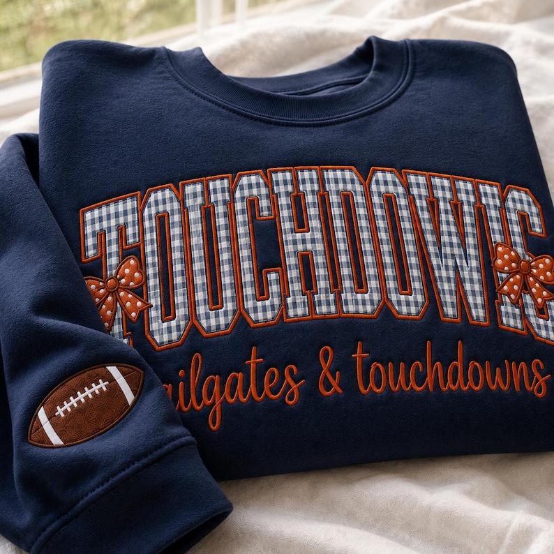 Op de afbeelding: Een marineblauw sweatshirt met ronde hals met het woord TOUCHDOWNS in een blauw-wit gingham patroon met oranje omlijningen. Twee oranje stippen strikken flankeren de tekst. Daaronder is de zin tailgates & touchdowns in het oranje geborduurd.