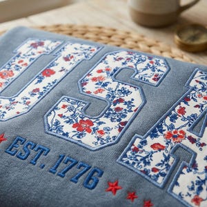 May include: A blue sweatshirt with the word "USA" in large, floral-patterned letters. The text "EST. 1776" is embroidered below, along with red star accents. The floral pattern features red and blue flowers.