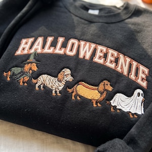 May include: A black sweatshirt featuring the embroidered text HALLOWEENIE in a curved font. Below the text are four dachshunds in costumes: one as a witch, one as a mummy, one as a hot dog, and one as a ghost, all rendered in detailed embroidery.