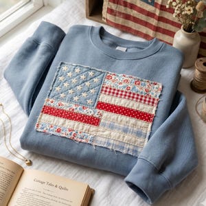 May include: A light blue sweatshirt with a patchwork American flag design. The flag features white stars on a blue field and red and white striped sections. The sweatshirt has a crew neck and ribbed cuffs.