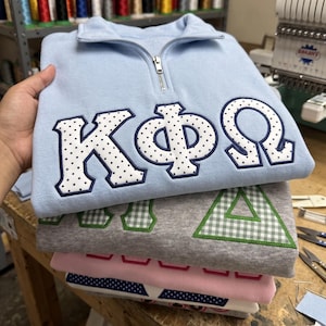 May include: A light blue quarter-zip sweatshirt featuring white Greek letters with a navy blue polka dot pattern and navy blue trim. Beneath it, a stack of folded sweatshirts in grey with green gingham letters, pink, and white with navy dots.