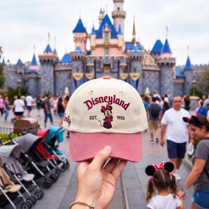 May include: A cream-colored baseball cap with a pink brim, featuring the embroidered text Disneyland EST. 1955 and a graphic of Minnie Mouse in the center. The name Emily is embroidered in cursive on the side of the cap in a dark pink thread.