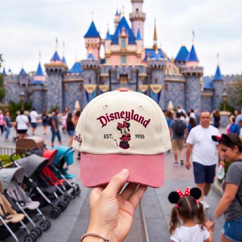 May include: A cream-colored baseball cap with a pink brim, featuring the embroidered text Disneyland EST. 1955 and a graphic of Minnie Mouse in the center. The name Emily is embroidered in cursive on the side of the cap in a dark pink thread.