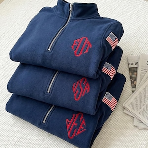 Könnte beinhalten: Drei marineblaue Fleece-Pullover gestapelt, jeder mit einem roten Monogramm und einem kleinen amerikanischen Flaggenaufnäher. Die Pullover haben einen Viertel-Reißverschluss und einen hohen Kragen. Die Artikel sind ordentlich auf einer weißen Oberfläche gefaltet.