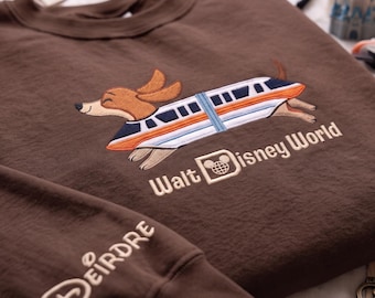 May include: A brown long-sleeve sweatshirt featuring an embroidered graphic of a dachshund running alongside a monorail train. The text Walt Disney World is embroidered below the graphic, and the name Deirdre is embroidered on the sleeve.