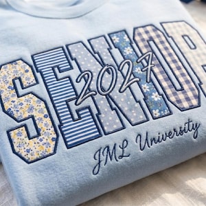 May include: A light blue sweatshirt featuring the word SENIOR in large block letters with varied blue and white patterns including floral, striped, polka dot, and gingham prints. The number 2027 and the text JML University are embroidered below.