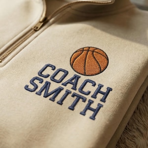 May include: A beige sweatshirt with a gold zipper and embroidered text and graphic. The text reads "COACH SMITH" in navy blue, with a basketball graphic above. The sweatshirt is folded.