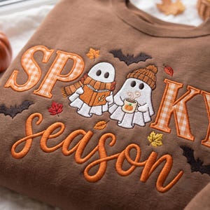 May include: A brown sweatshirt featuring embroidered text that reads SPOOKY season. The design includes two white ghosts, one wearing a scarf and reading a book, the other wearing a beanie and holding a mug with a pumpkin, surrounded by bats and leaves.