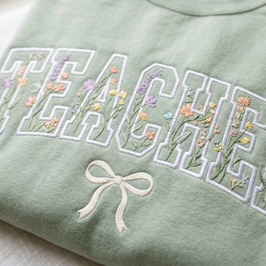 May include: A sage green sweatshirt featuring the word "TEACHER" in white embroidered block letters, filled with colourful floral embroidery. A white bow is embroidered below the text.