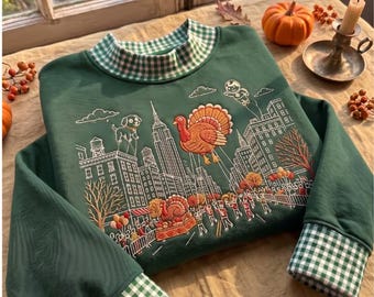Op de afbeelding: Een donkergroen sweatshirt met een groen-witte gingham kraag en boorden. De voorkant toont een geborduurde scène van een stadsoptocht met een grote kalkoenballon, een hondenballon, een astronautenballon, gebouwen en marcherende figuren.