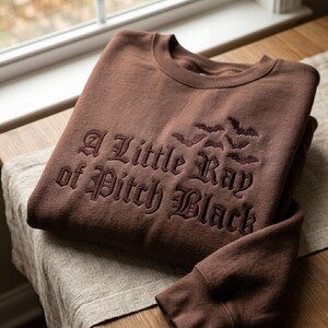 May include: A brown sweatshirt with the embroidered phrase "A Little Ray of Pitch Black" in a gothic font, along with a bat graphic. The sweatshirt is folded on a wooden surface.
