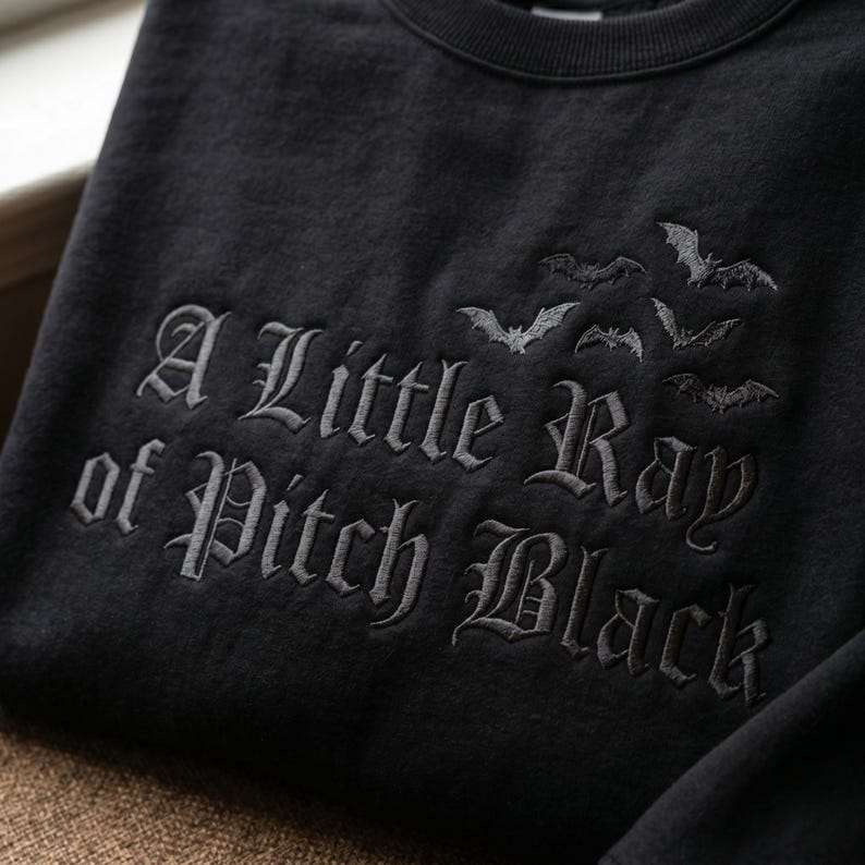 May include: Black sweatshirt with the embroidered phrase "A Little Ray of Pitch Black" in gothic lettering, accompanied by a flock of bats. The dark design is perfect for Halloween or gothic-themed apparel.