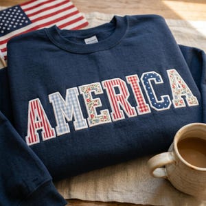May include: A navy blue sweatshirt displaying the word "AMERICA" in a patchwork design. The letters incorporate red and white stripes, blue gingham, floral patterns, and stars. An American flag and a mug of coffee are visible.