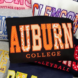 Può includere: Una felpa nera con la parola AUBURN in grandi lettere in stile blocco con motivi a pois e floreali arancioni, seguita dalla parola COLLEGE in lettere arancioni più piccole. Il design è applicato con un bordo cucito visibile.