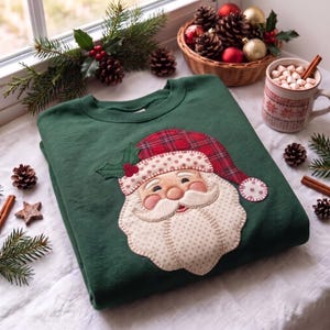 May include: A dark green crewneck sweatshirt featuring a large, centered fabric applique of a Santa Claus face. The Santa design includes a red plaid hat, a cream-colored beard with a subtle polka dot pattern, and green holly leaves.