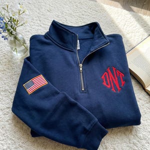 Op de afbeelding: Een marineblauw sweatshirt met een kwartrits, zilveren rits en een kleine Amerikaanse vlagpatch op de linkermouw. De rechterborst heeft een rood geborduurd monogram. Het sweatshirt is op een witte, getextureerde ondergrond gevouwen.