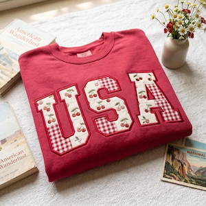 Pode incluir: Um moletom vermelho com a palavra "USA" aplicada na frente. As letras são brancas com um padrão de cerejas e detalhes em xadrez vermelho. O moletom está em uma superfície branca com livros e um vaso de flores.