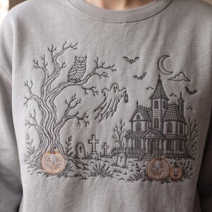 May include: A light gray crewneck sweatshirt featuring black line-art embroidery of a haunted house, a ghost holding a lantern, an owl in a tree, and tombstones. The design includes three orange-stitched jack-o'-lanterns and a crescent moon.