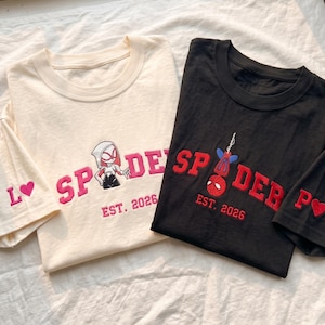 May include: A cream t-shirt with pink embroidered text reading SPIDER and a character graphic, and a black t-shirt with red embroidered text reading SPIDER and a hanging character graphic. Both shirts feature EST. 2026 and a heart on the sleeve.
