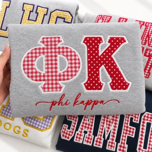 Könnte beinhalten: Ein graumeliertes Sweatshirt mit großen griechischen Buchstaben Phi und Kappa, die in rot-weißen Mustern appliziert sind. Das Phi ist in rot-weißem Vichy-Karo gehalten, während das Kappa ein rot-weißes Punktemuster aufweist. Darunter ist der Text phi kappa in roter Schreibschrift gestickt.