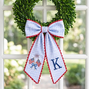May include: A green wreath with a large blue and white gingham bow. The bow has red scalloped edges and features embroidered American flags, fireworks, and the letter "K". The wreath is hanging on a white window.