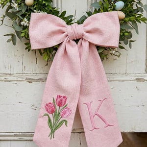 May include: A large pink bow with long streamers, embroidered with pink tulips and the letter "K". The bow is part of a wreath with greenery and small white flowers, and decorative eggs. The bow is made of a textured fabric.