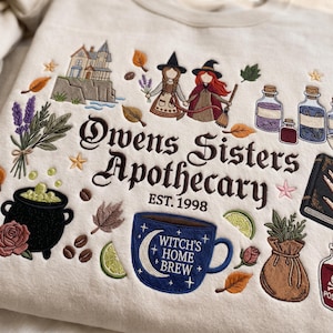 Op de afbeelding: Een beige sweatshirt met borduursels van een huis op een klif, twee figuren met hoeden, toverdrankflesjes, een zwarte ketel, een bruine buidel en een blauwe mok met de tekst "WITCH'S HOME BREW". De centrale tekst luidt "Owens Sisters Apothecary EST. 1998".