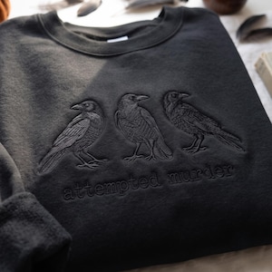 Op de afbeelding: Een zwart sweatshirt met ronde hals en een ton-sur-ton borduursel van drie kraaien die op een rij staan. Onder de vogels is de tekst "attempted murder" geborduurd met bijpassend zwart garen, wat zorgt voor een subtiel en gestructureerd effect.
