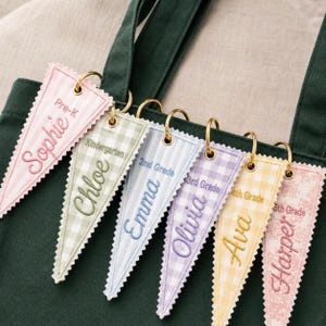 May include: Six triangular fabric pennants with scalloped edges attached to a dark green bag by gold metal rings. Each pennant features embroidered names and grade levels: Sophie Pre-K, Chloe Kindergarten, Emma 2nd Grade, Olivia 3rd Grade, Ava 4th Grade, and Harper 5th Grade.