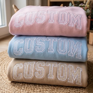 Könnte beinhalten: Ein Stapel von drei Sweatshirts in Pastellfarben: Rosa, Hellblau und Beige. Jedes Sweatshirt hat das Wort "CUSTOM" in großen, weißen Buchstaben im College-Stil mit einem Vichy-Muster. Die Sweatshirts sind sauber gefaltet.