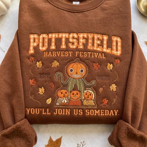 Puede incluir: Una sudadera marrón de cuello redondo con texto bordado que dice POTTSFIELD HARVEST FESTIVAL EST. 1502 YOU'LL JOIN US SOMEDAY. El diseño incluye una figura central con cabeza de calabaza, tres personajes de calabaza más pequeños y hojas dispersas.