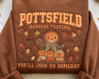 May include: A brown crewneck sweatshirt featuring embroidered text that reads POTTSFIELD HARVEST FESTIVAL EST. 1502 YOU'LL JOIN US SOMEDAY. The design includes a central pumpkin-headed figure, three smaller pumpkin characters, and scattered orange and brown leaves.